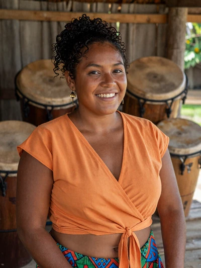 Karina, 28, Belizean Garifuna woman from Hopkins with joyful smile in orange top near traditional drumming hut - Latin 312 verified profile